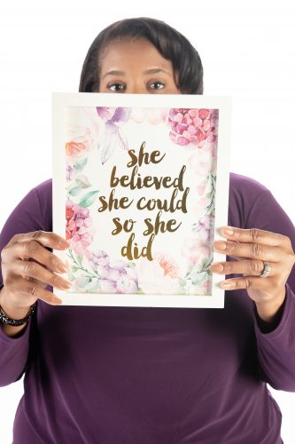 Karen Bell Morgan holds a framed print reading “She believed she could, so she did” in front of her face, with only her eyes visible above it.
