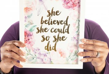 Karen Bell Morgan holds a framed print reading “She believed she could, so she did” in front of her face, with only her eyes visible above it.
