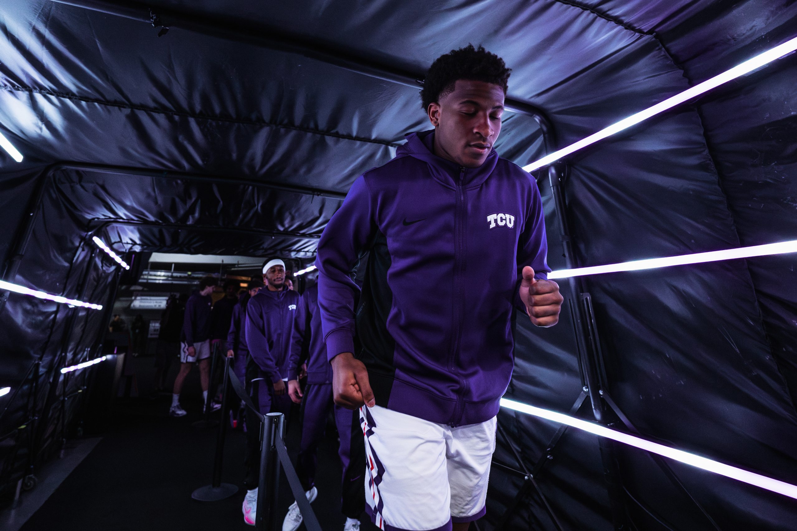 Xavier Edmonds, in a purple TCU zip-up and white shorts, walks through a tunnel lit with white light bars as his teammates line up behind him.