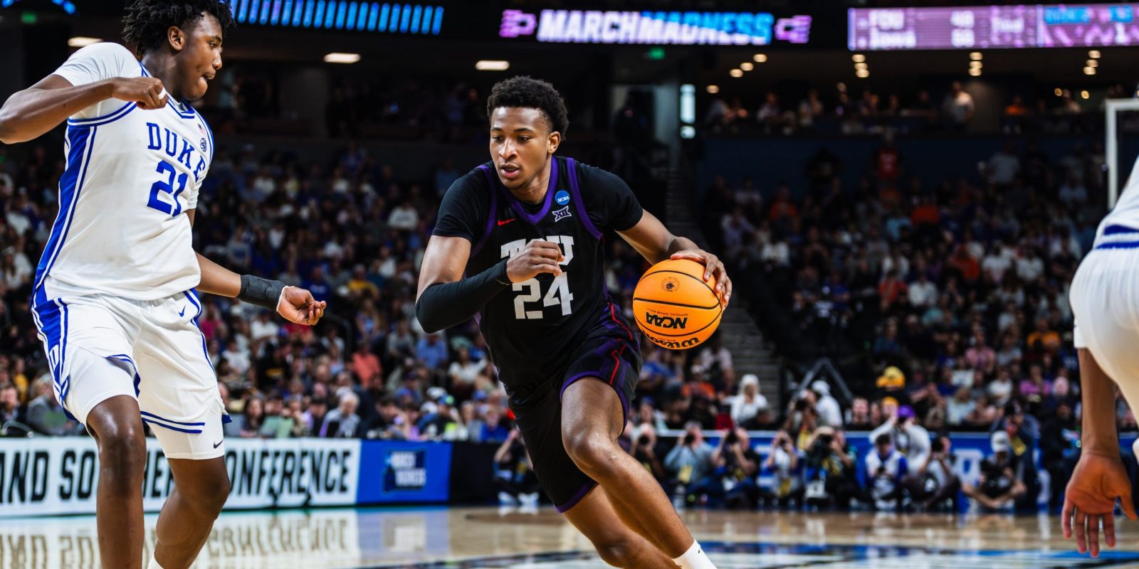 Xavier Edmonds, in a black TCU No. 24 jersey, drives with the ball past a defender in a white Duke uniform in front of a packed arena.