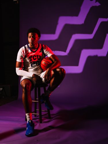 Xavier Edmonds sits on a wooden stool in a white TCU No. 24 jersey and blue sneakers, holding a basketball against his knee, in front of a purple backdrop lit with zigzag shapes.