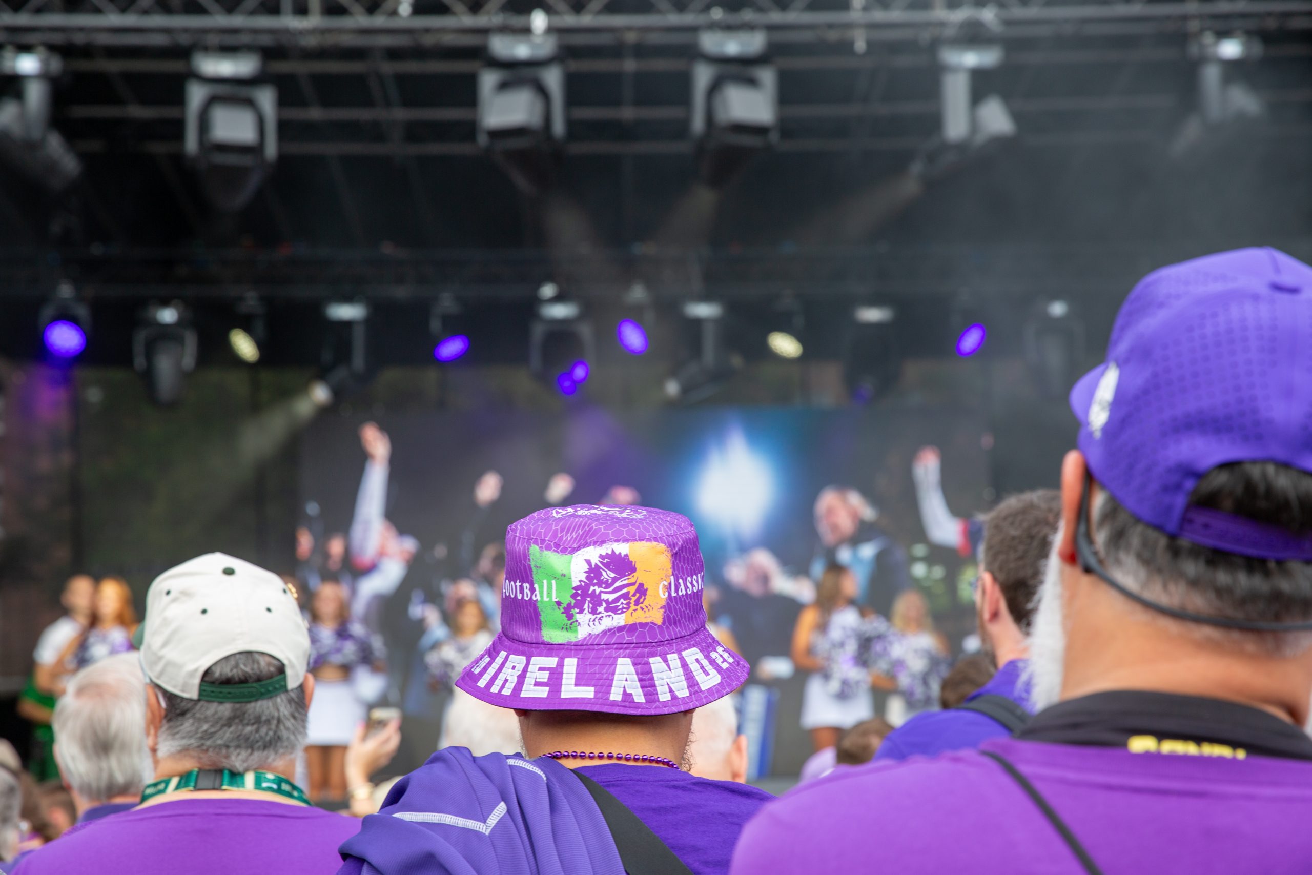 A crowd dressed in purple watches performers with pom-poms on a lit stage. One person wears a purple bucket hat that reads "Football Classic" and "Ireland 26" and has a horned frog logo.