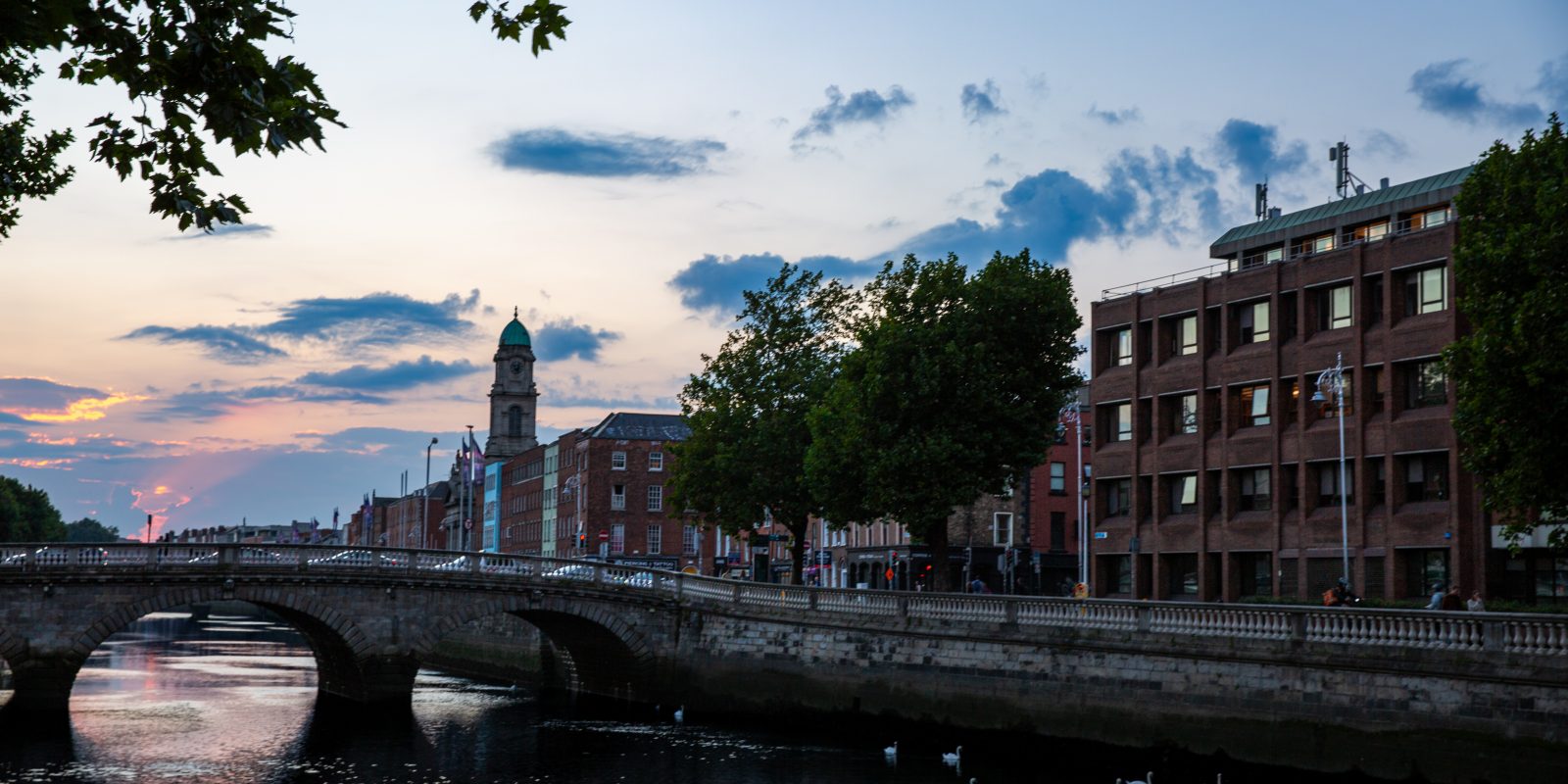 A stone bridge spans a river at dusk, with brick buildings and a distant clock tower along the bank under a pink and blue sky.