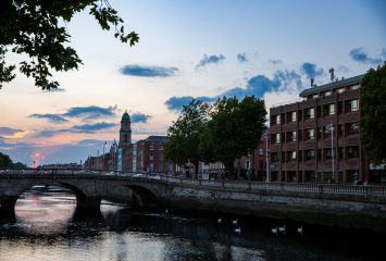 A stone bridge spans a river at dusk, with brick buildings and a distant clock tower along the bank under a pink and blue sky.
