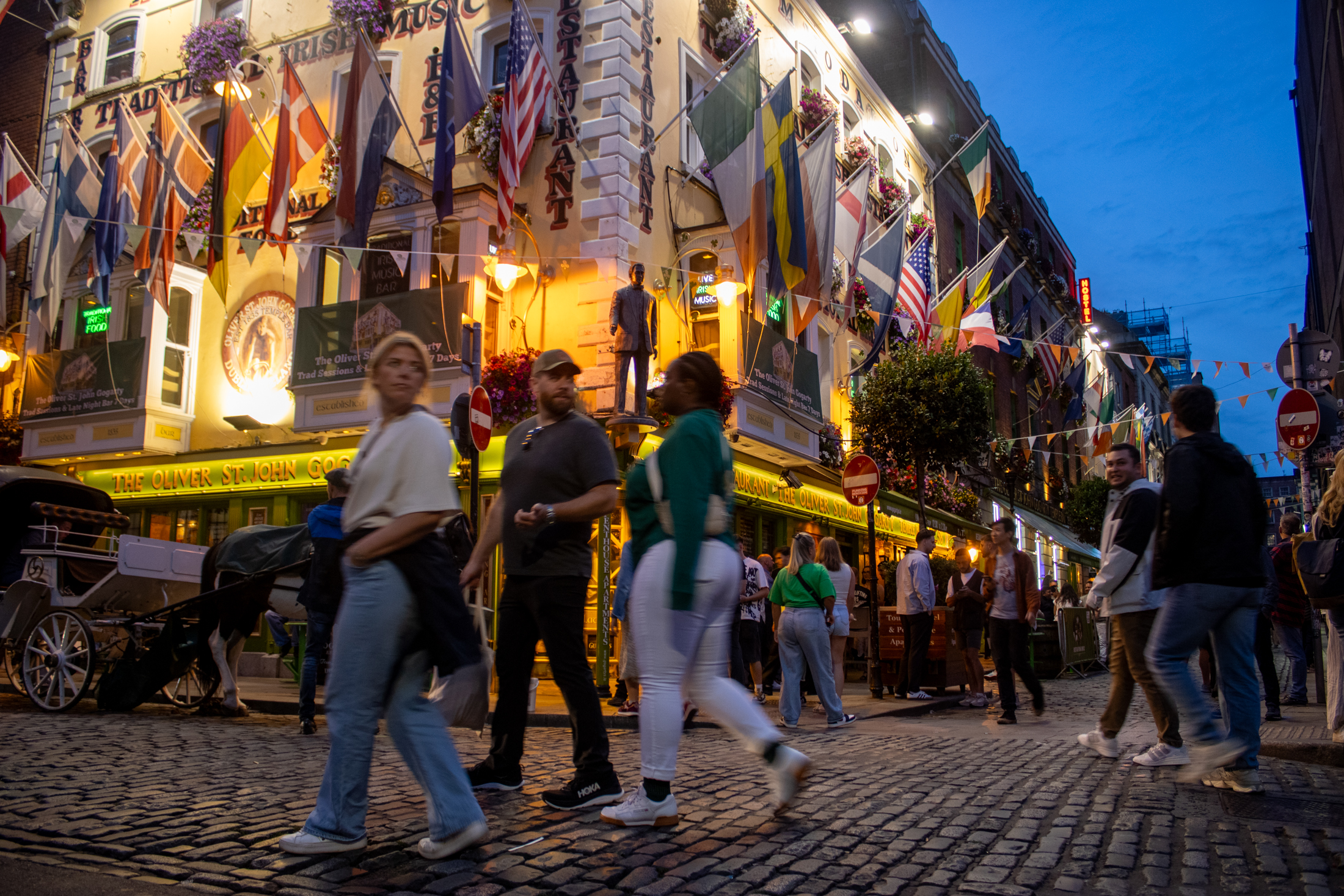 People stroll along a cobblestone street at dusk past The Oliver St. John Gogarty pub, hung with international flags and flower baskets. A horse-drawn carriage waits at left.