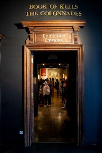 Gold letters reading "Book of Kells / The Colonnades" sit above a carved wooden doorway marked "Exhibition Entrance," with visitors visible inside.