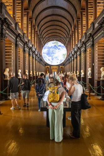 Visitors fill Trinity College Dublin's Old Library, with towering wooden bookshelves beneath an arched ceiling and a large illuminated globe hanging overhead.