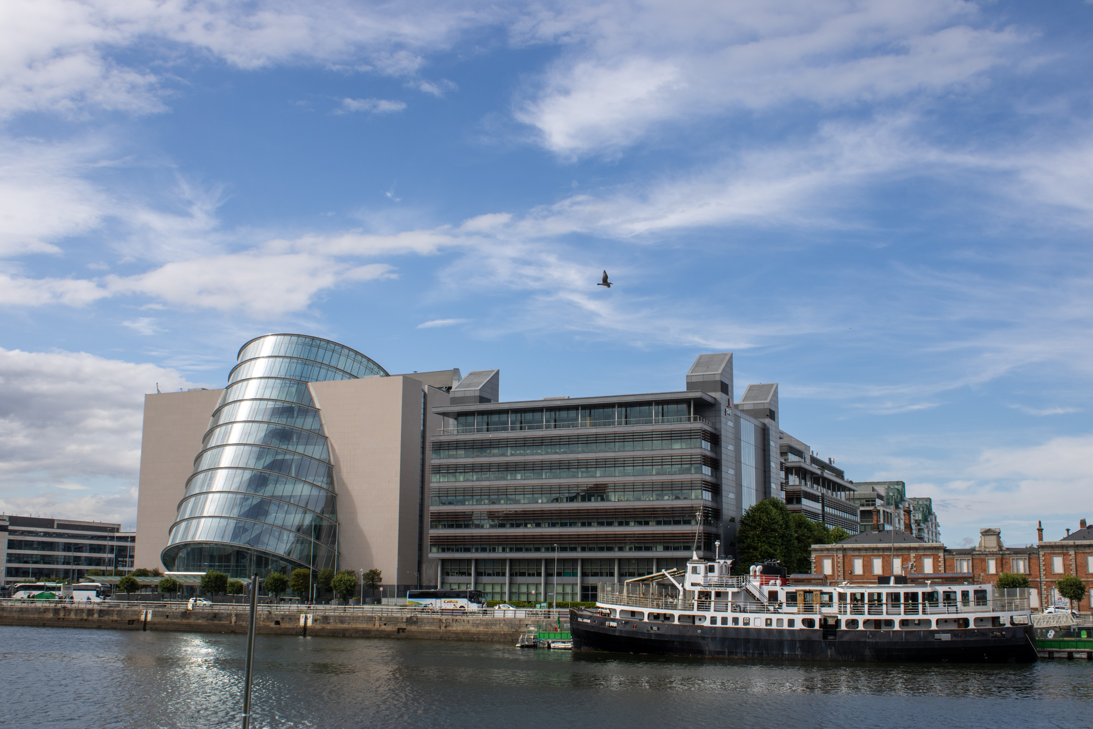 A modern glass building with a tilted, cylindrical front stands beside other multistory buildings along a river in Dublin, with a black-and-white boat docked along the bank and a bird flying overhead under a partly cloudy sky.