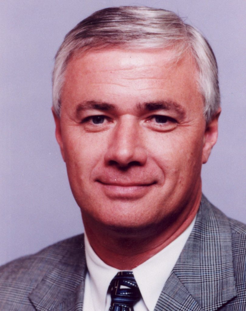Dennis Franchione, in a gray plaid jacket, white shirt and patterned tie, poses for a portrait against a lavender backdrop.