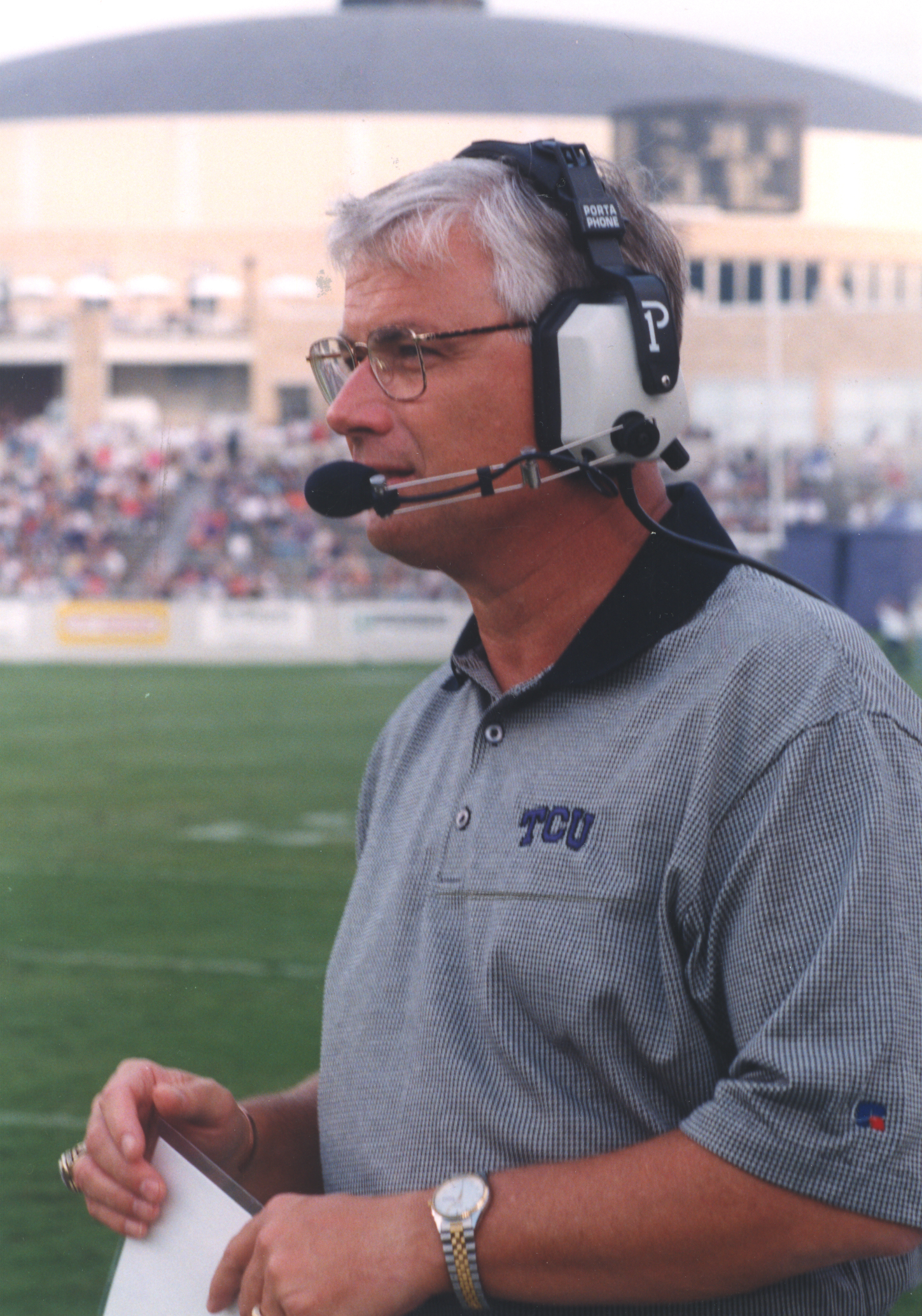 Dennis Franchione, wearing a headset and a gray TCU polo, stands on the sideline with a stadium crowd behind him.