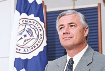 Dennis Franchione, in a gray suit and tie, stands beside a flag bearing the TCU seal.