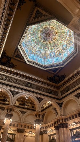 A colorful octagonal stained-glass dome tops an ornate hall at Teatro Colón in Buenos Aires, with carved decorative trim, arches and columns topped with lights below.