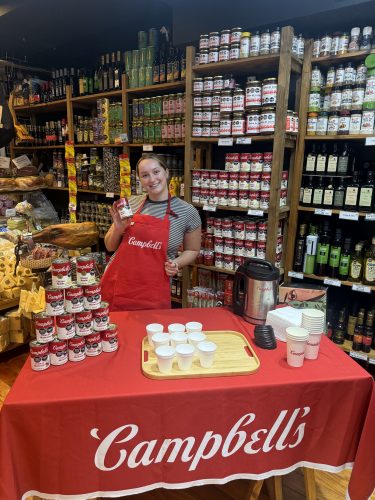 Kierstyn Wright, wearing a red Campbell’s apron, smiles and holds up a can of soup behind a Campbell’s soup tasting table with stacked cans and sample cups, in a store lined with shelves of jars, spices and bottles.