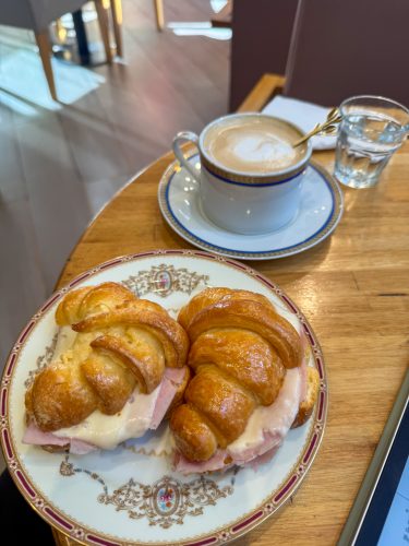 Two medialunas filled with sliced meat and cheese sit on a decorative plate on a wooden cafe table, next to a cup of coffee with latte art and a glass of water.
