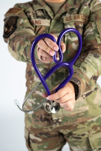 Hands hold a purple stethoscope bent into a heart shape in front of a camouflage uniform with a “U.S. Air Force” tape on the chest.