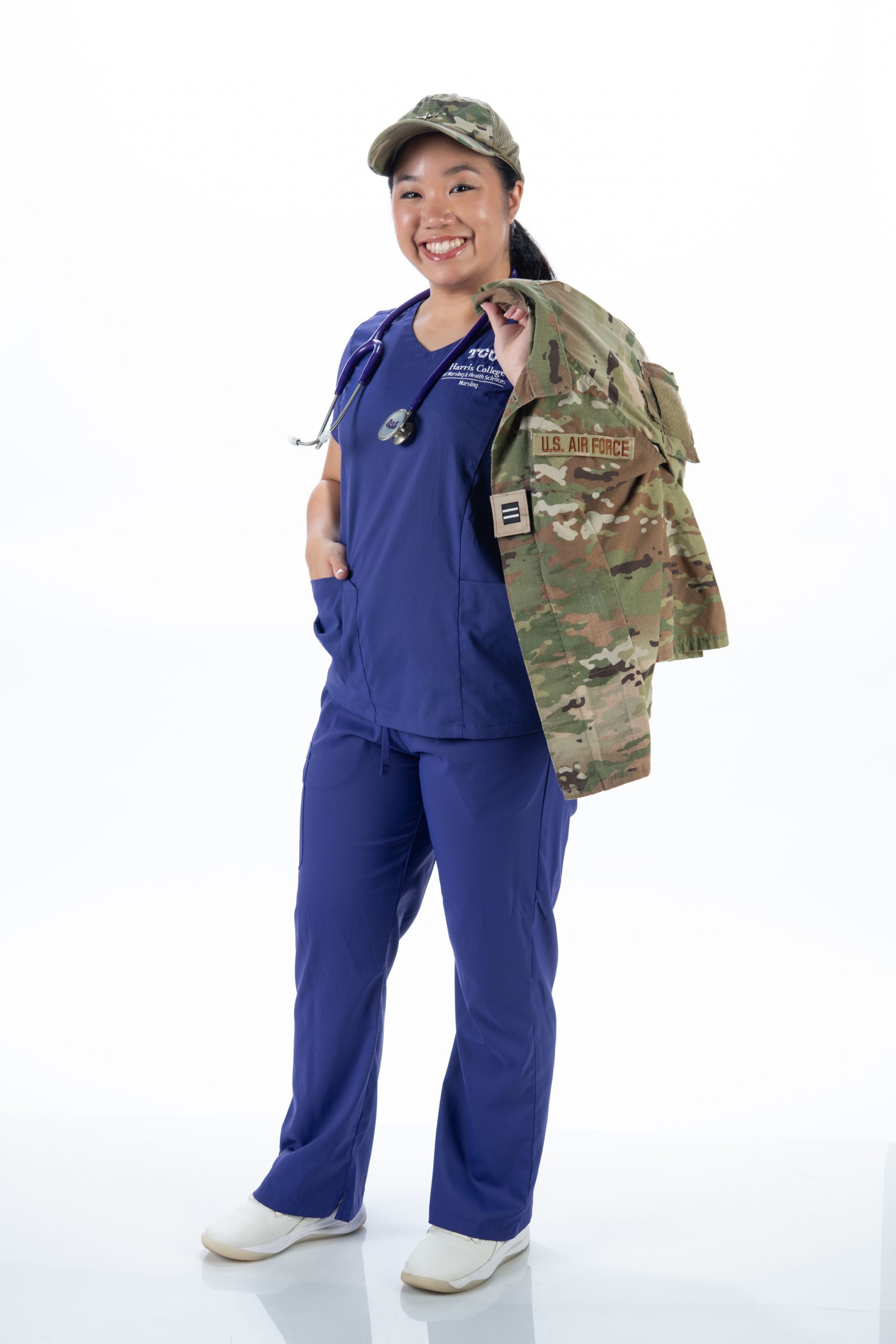 Fayette Jiang smiles in purple TCU Harris College of Nursing & Health Sciences scrubs and a camouflage cap, a stethoscope around her neck and a U.S. Air Force uniform jacket over her shoulder.